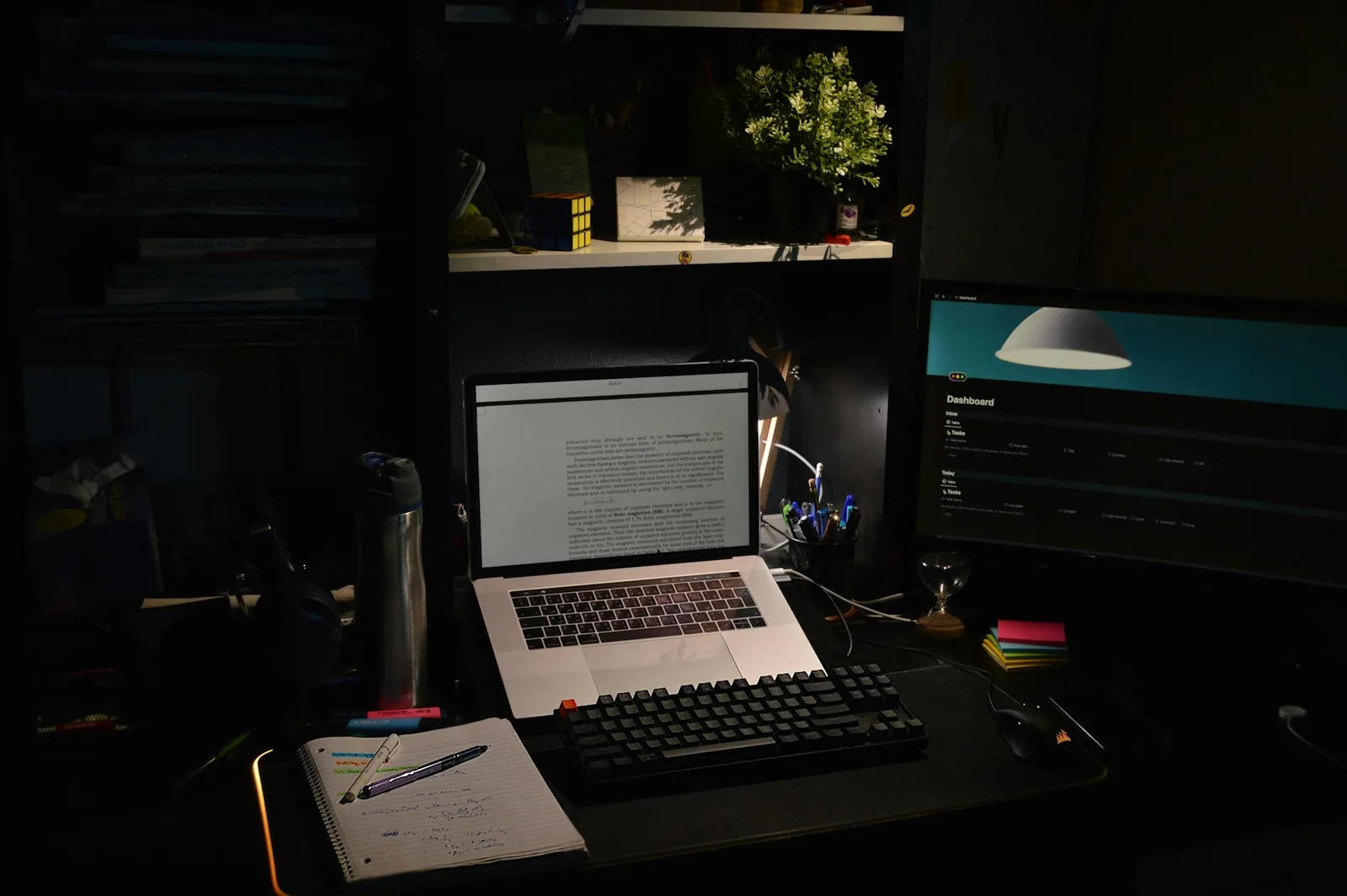 Dark desk at night with a laptop open beside a notebook and pens