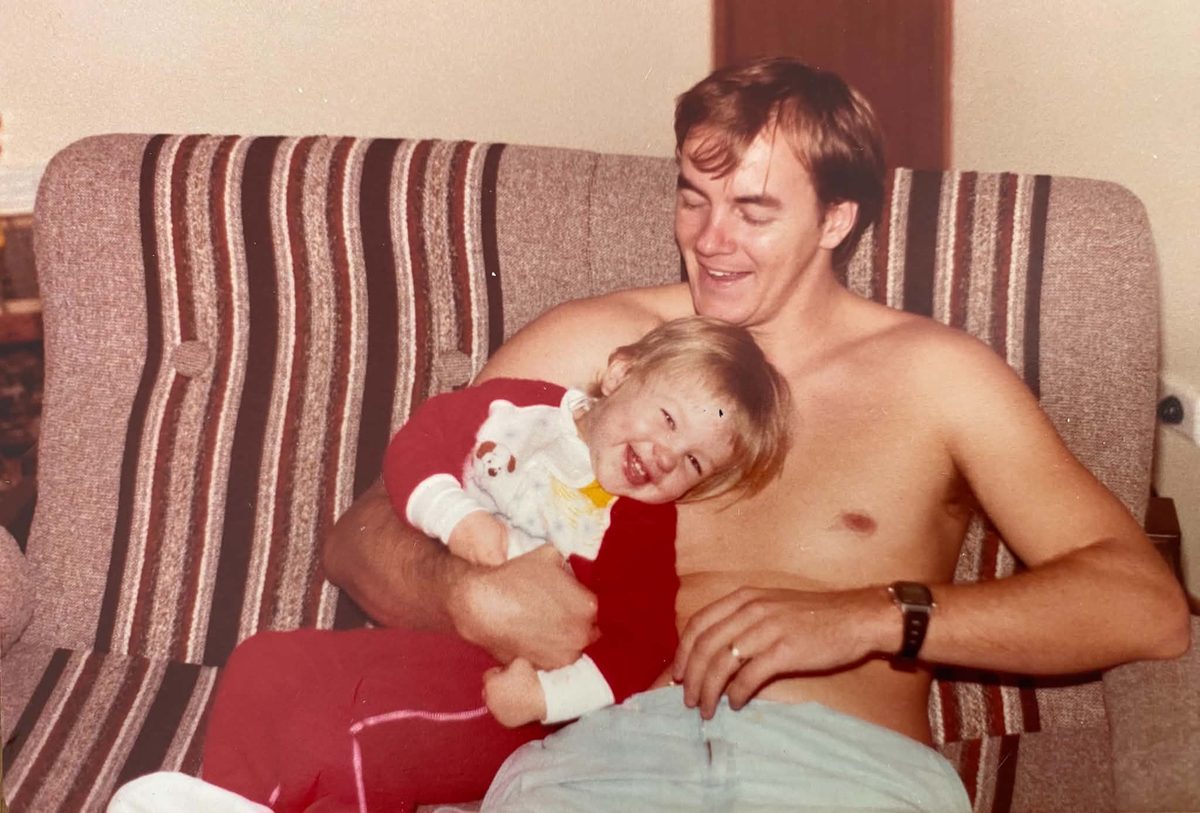 Mark Rasmussen in the mid 1980s, laughing on a striped couch with his baby daughter Hayley in his arms